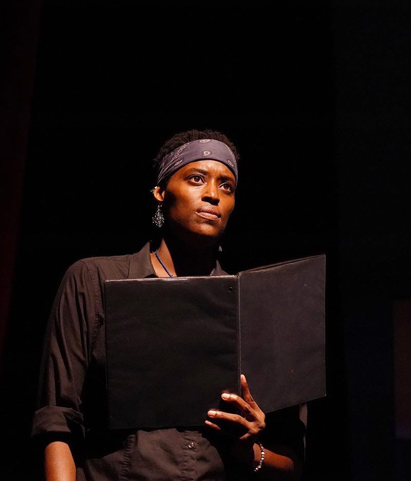 black female reading from a black book with blue headband on staring out at audience